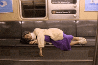 Video gif. Exhausted grandma wearing a neck brace lies on a subway seat with her head resting on a purse, snoozing before being startled awake.