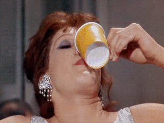 Movie gif. A drunk Mag Wildwood as Dorothy in Breakfast at Tiffany's wobbles and tosses back the contents of a yellow cup, looking bleary-eyed at someone offscreen. She's all dressed up but barely holding it together, wearing gaudy diamond earrings, heavy eye makeup, and hair swept back in an elegant updo.