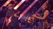 ACM Awards gif. Two people. Man in tan cowboy hat stums guitar while singing into microphone as his head turns from facing Dua Lipa to the mic, Dua Lipa takes hand off mic its stand while continuing to sing with a happy expression as she sways from side-to-side.