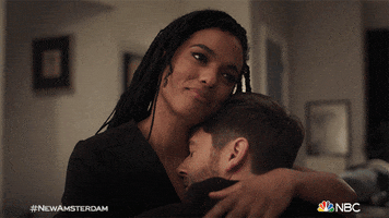 TV gif. Ryan Eggold as Dr. Dan and Freema Agyeman as Dr. Helen are hugging closely on a couch. He squishes closer to her and she rubs his back, with a soft smile on her face. 