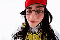 Music Video gif. From Billie Eilish's music video for "LUNCH", Billie is on a stark white background dressed in a long sleeve white shirt with a yellow and black jersey over the top, with a red hat on. She starts very close to the camera with her eyes wide, and backs up while clapping after.