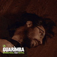 Video gif. A man and woman lay prostrate on a hardwood floor under creepy flickering lights. They each grab their chests and convulse as if attacked by an unseen force. 