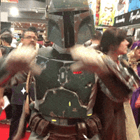 Video gif. A Boba Fett cosplayer forms a heart at us with his hands and tilts his head in a cute, loving way. 