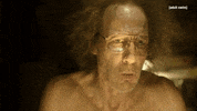 TV gif. A scene from Dream Corp LLC. A man with gray frizzy hair and aviator glasses stands without his shirt on. He has a surprised and relieved expression on his face. He says, “We're back.”