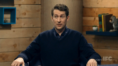 TV gif. Scott Aukerman on Comedy Bang Bang sits in a chair with his arms out, looking to the side and then looking forward at us with an excited grin as he says "yikes," which appears as text.