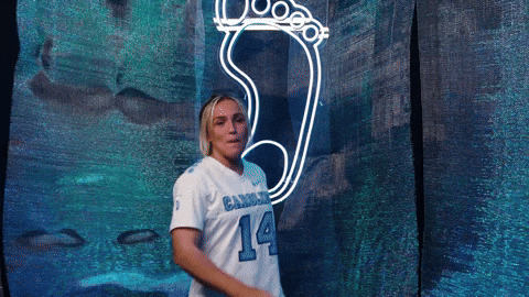 Sports gif. Livi Lawton, a lacrosse player from the University of North Carolina, dances in front of a neon sign that looks a footprint.