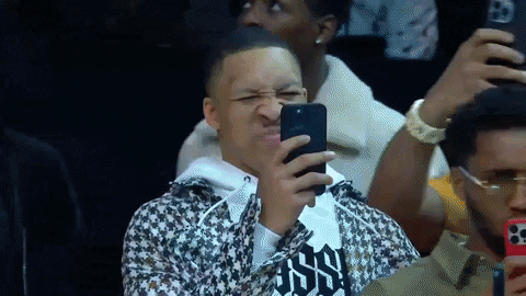 Video gif. Donovan Mitchell and Grant Williams are at the 2023 NBA All-Star Dunk Contest. Both are recording the dunks on their phones, scrunching their noses and looking frustrated or uncomfortable about what they're filming. 