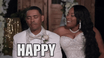 Reality TV gif. A bride and groom sit side by side from Bridezillas as the bride grins and leans close to the stone faced groom. Text, "Happy wife happy life." 