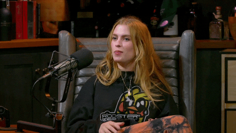 Video gif. Barbara Dunkelman on the Roosterteeth Podcast sits in a large chair in front of a microphone. She contorts her face into an over exaggerated sad expression and she yells out in fake agony, “I know!” and laughs at herself.