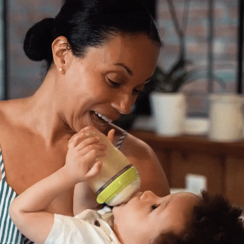 A woman smiling while feeding a baby from a green-capped bottle.