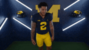 Sports gif. Blake Corum, a football player for the University of Michigan, stands in a Michigan Athletics-themed set wearing his jersey. He leans towards us, circling his hand and then holding it up to his ear like he's inviting you to whisper something to him.