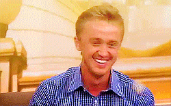 Celebrity gif. Tom Felton is being interviewed but something makes him laugh big time and he covers his mouth with both hands as he cracks up.