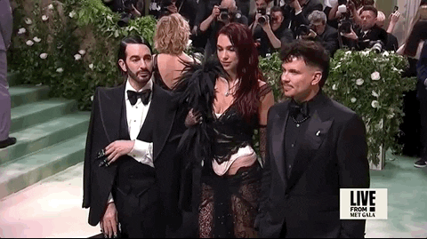 Met Gala 2024 gif. Dua Lipa is dressed in a black and white vampy boudoir-style custom Marc Jacobs two-piece with corsetry detailing, a feather boa, and black lace low-rise skirt. She's standing with Marc Jacobs and internet personality Char DeFrancesco.