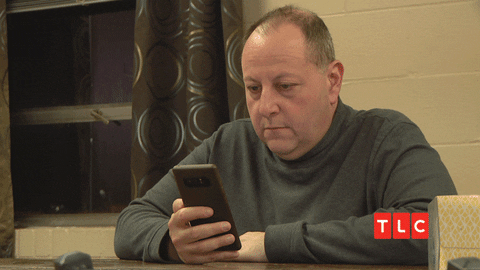 Reality TV gif. David from 90 Day Fiancé sits at a table holding his phone, looking straight-faced but surprised, raising his eyebrows and glancing side to side.