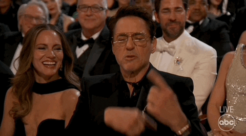 Oscars 2024 GIF. Robert Downey Junior, seated at the Oscars, looks at us and nods with a sarcastic smirk, gesturing the sign for rolling, saying “keep it going.”