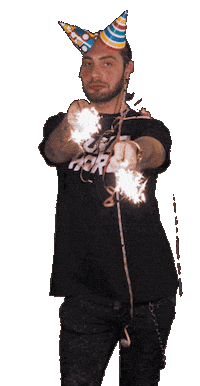 Sticker gif. Tomy Kotty, draped with ribbons and wearing two party hats resembling horns, sways his hips while waving lit sparklers at us.