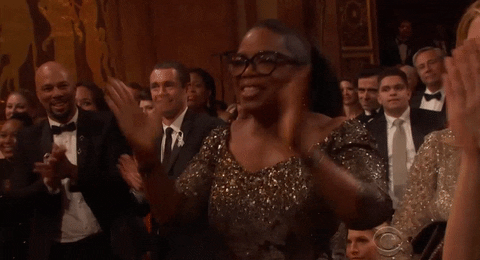 Celebrity gif. Oprah Winfrey stands in the crowd at the Tony Awards wearing a sleek, glittering gray dress and a pair of black spectacles. She claps approvingly and exclaims "Yes!" passionately.