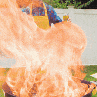 Video gif. Man wearing a yellow apron and holding a spatula stands behind a round outdoor grill. Flames burst from the grill into the air. 