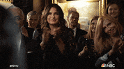 TV gif. Mariska Hargitay as Olivia Benson on Law and Order SVU claps and smiles, somewhat reserved, among a group of older people smiling and applauding.