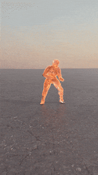 Digital art gif. A person stands on desolate ground fully engulfed in flames as they begin stumbling sideways. The flames lag behind as the person eventually falls to their side on the ground. 