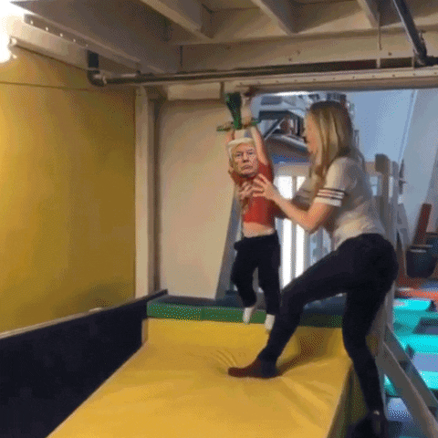 Meme gif. Donald Trump's face superimposed onto a small child, mounted onto a zipline by his mother, then released, as he dangles haplessly, zipping across a foam pit and, smack, directly into a giant mat labeled "all of us."