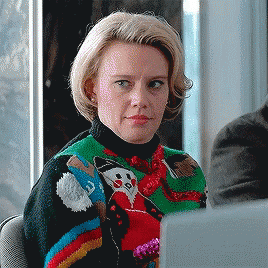 Celebrity gif. Kate McKinnon is wearing an ugly Christmas sweater while sitting in what appears to be a meeting. She looks to her side and slowly winks awkwardly while smiling at someone.