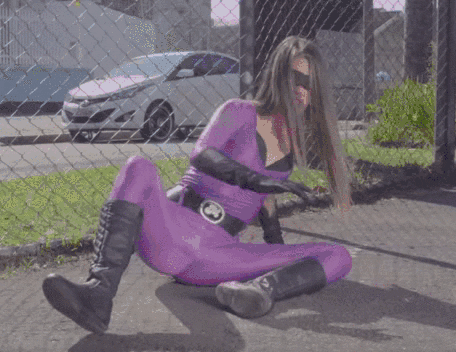 Video gif. Woman in a spandex superhero suit and black mask sits on the ground with her knees spread, clenching with all of her strength until a bright purple glowing light forms in her crotch, as she screams powerfully.