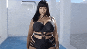 Video gif. A curvy fashion model, wearing a black bodysuit with cutouts along her hips and obliques, smizes at us and maintains a power pose.  