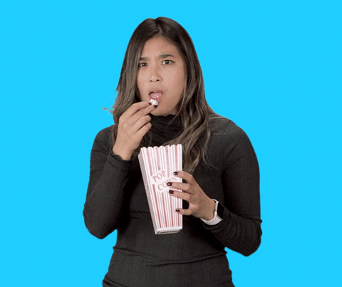 Video gif. Holding a popcorn container and tossing kernels into her mouth, a woman nods agreeably.