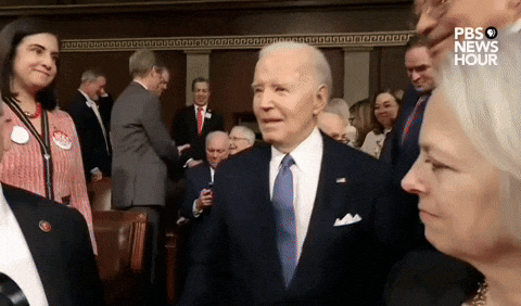 Video gif. President Joe Biden enters a crowded House of Representatives for his 2024 State of the Union Address. As he makes his way down the crowded aisle, he spots someone in the distance and stops in his tracks, looking amused. The camera pans over and shows a smug Representative Marjorie Taylor Green in the crowd wearing a red Make American Great Again hat. 