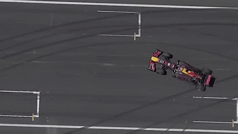 Video gif. Drone footage looks down on a formula one car making donuts on a blacktop. Smoke billows out from the back tires.