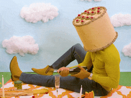 Video gif. A man with a birthday cake for a head, sits on a picnic blanket with a fancy, romantic meal spread out for us. The backdrop is construction paper grass, sky, and cotton-ball clouds. He opens a bottle of champagne which sprays everywhere before holding his hand out in presentation. Text, "Happy anniversary."