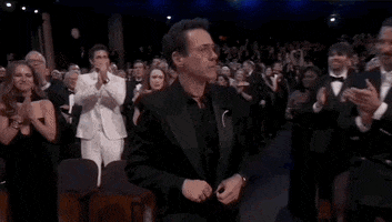 Oscars 2024 gif. Robert Downey Jr stands to receive the award for Best Supporting Actor. While buttoning up his blazer, he quickly turns to face the standing crowd, who applauds him while he waves out to them. He turns back around with a serious expression spread across his face. 