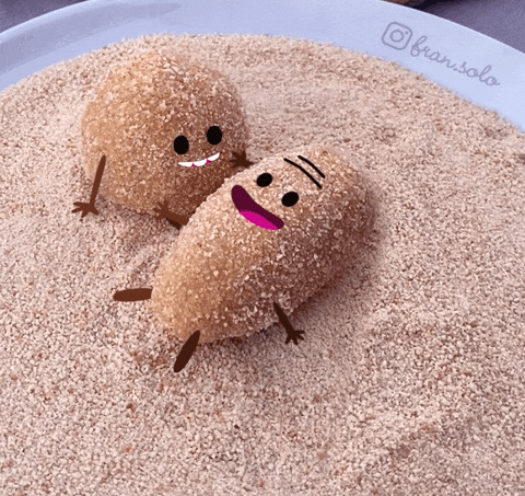 Video gif. In a bowl full of cinnamon sugar, two croquettes with adorable faces smile. One rolls excitedly back and forth in the sugar.