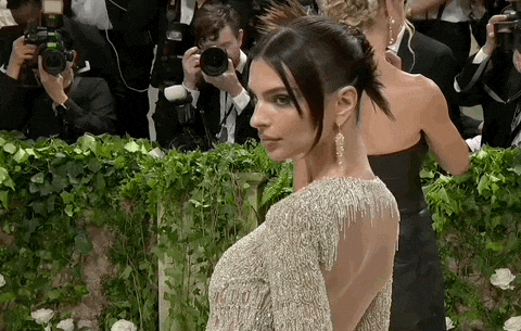 Met Gala 2024 gif. Emily Ratajkowski shows the upper back and front of her long sleeve pale gold Versace gown with a crew neckline. Her hair is swept back into a tight bun with three ends flaring out resembling bird feathers. Two short loose strands hang down and frame her face.