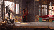 TV gif. Jennifer Metcalfe as Mercedes and Chelsee Healey as Goldie from Hollyoaks burst into a room announcing their return, carrying luggage and smiling. 