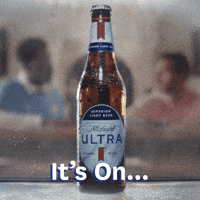 Sponsored gif. Bottle of Michelob ULTRA comes into focus before blurring. Two men are shown behind it at a bar. They stare intently at each other. Text reads, “It’s on…”