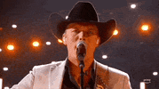 ACM Awards gif. Male musician in cowboy hat strums guitar out of frame while singing happily into a standing mic as the camera rotates around the singer, they wink and smile into the camera.