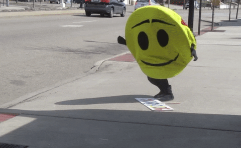 Video gif. A yellow smiley faced mascot slowly falls to their knees to try to pick up the sign that’s on the sidewalk. The mascot loses their balance and falls backwards. 