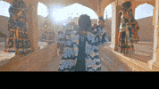 Music video gif. Badshah, wearing thick sunglasses, lifts the hood of his coat over his head in slow motion. Video then cuts to him standing with a woman singing while Badshah points at us. Text reads, “ITS YOUR BOY BADSHAH.”