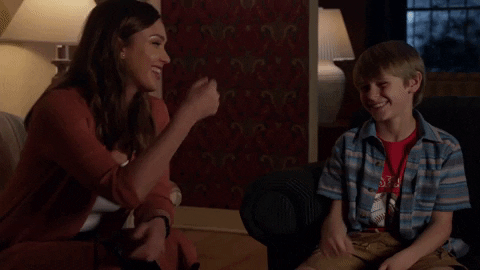 TV gif. Jessica Alba as Nancy McKenna in La’s Finest leans over and fist bumps a young boy sitting next to her.