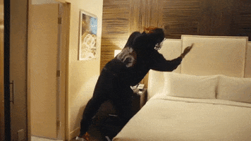 Video gif. A man with long hair launches himself onto a bed and lands on his back with arms spread wide.
