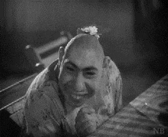Movie gif. Schlitzie, an actor with microcephaly, in "Freaks" sits at a table, wearing a dress, doubling over in laughter and covering his face.