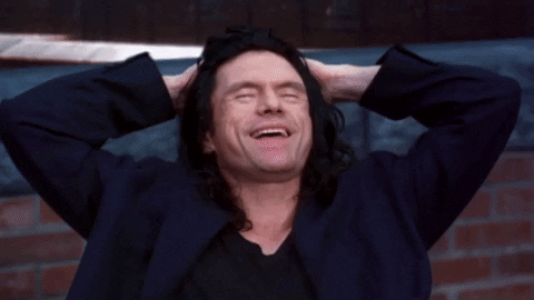 Movie gif. Tommy Wiseau as Johnny in The Room has his hands behind his head as he laughs and then he stands up, replying, "What a story, Mark!"