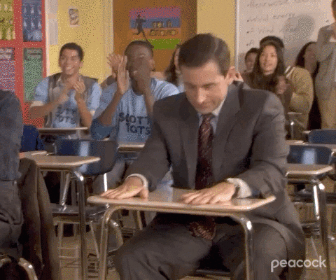 The Office gif. Steve Carell as Michael sits in a classroom among cheering students as he drops his head and gives a forceful double thumbs up. 