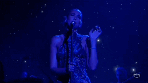 ACM Awards gif. While gripping mic stand with one hand Dua Lipa waves free hand at ear-level height before bringing it down then back up again as she sings and lightly shakes her head from side-to-side.