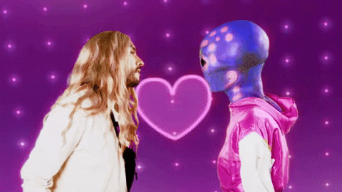 Video gif. A man and a person dressed as a gray alien with a pink hoodie slowly lean in towards each other to kiss as a flashing, neon heart grows and stars burst in the background. 