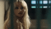Music video gif from Sabrina Carpenter's music video for "Please Please Please." She sits in a jail cell behind bars, smiling and scrunching up her face, then stands up.