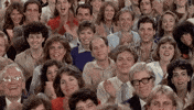 Video gif. Crowd of people who look like they’re from the 1980s looks at us and claps all in unison, and smile.