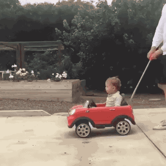 Video gif. A toddler getting slowly pushed in a manual toy car quickly turns around when he sees a bigger, automatic toy car pull up. The driver of the automatic car, a swagged out pug with sunglasses and a jersey on, pulls up next to the toddler to pause before jetting off again. 